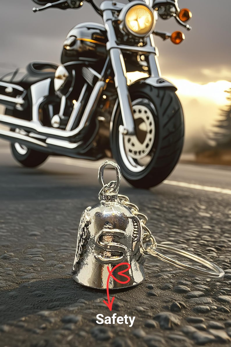 May include: A silver motorcycle bell with a keychain, with the word "Safety" in the image. A black and chrome motorcycle is in the background. The bell is a common accessory for motorcycle riders.
