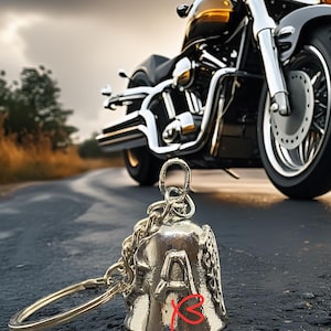 May include: A silver motorcycle bell with the word "Angel" and the letter "A" on the side. The bell is attached to a chain and a ring. A black and gold motorcycle is in the background.