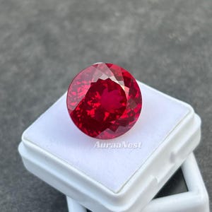 May include: A faceted, round, deep red gemstone. The gem is set on a white plastic base. The image is well-lit, showcasing the gem's brilliance. The text "AuraaNest" is visible on the base.