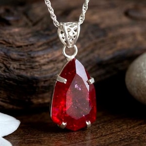May include: A teardrop-shaped red gemstone pendant set in a silver prong setting, suspended from a silver chain. The faceted stone displays a deep crimson hue with internal reflections, held by a decorative silver bail with a lattice pattern.