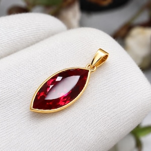 Puede incluir: Un colgante con una piedra preciosa de color rojo intenso en talla marquesa, engastada en un bisel de metal dorado pulido. El colgante presenta una superficie facetada que refleja la luz y una anilla de metal dorado para colgarlo de una cadena.