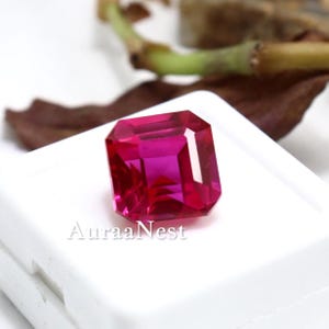 May include: A vibrant, square-cut, faceted gemstone in a deep magenta-red hue, resting on a white display base. The stone exhibits sharp, geometric edges and internal light reflections. The text AuraaNest is printed in black on the white base.