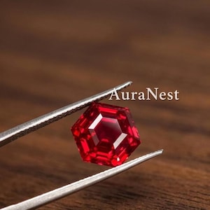 Könnte beinhalten: Ein leuchtend roter Edelstein mit Hexagonschliff, der von einer silberfarbenen Metallpinzette gehalten wird. Der facettierte Stein zeigt tiefe karmesinrote Farbtöne und scharfe geometrische Kanten. Der Text AuraNest ist oben rechts zu sehen.