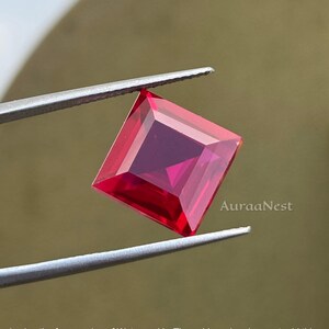 May include: A square-cut, vibrant red gemstone is held by silver tweezers. The gem has a deep, rich colour and a faceted surface, reflecting light. The text "AuraaNest" is visible in the lower right corner.