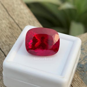 May include: A cushion-cut, faceted, deep red gemstone. The gem is set in a white display box, with the text "AuraaNest" printed on the box. The background is a blurred view of wood and greenery.