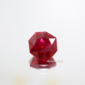 Può includere: Una gemma di rubino sfaccettata, taglio ottagonale. La gemma è di un colore rosso intenso e vibrante, con molteplici sfaccettature che riflettono la luce. La pietra preziosa è posta su uno sfondo bianco, con il suo riflesso visibile sotto.