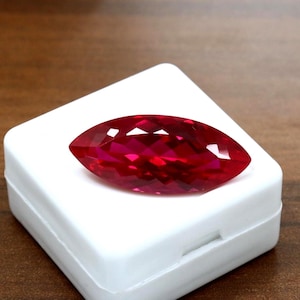 Puede incluir: Una piedra preciosa de corte marquesa con un color rojo intenso y vibrante y una superficie facetada que refleja la luz. La piedra está colocada sobre una pequeña caja de presentación cuadrada de plástico blanco con esquinas redondeadas.