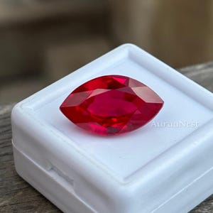 May include: A faceted, marquise-cut ruby gemstone, displayed in a white plastic case. The ruby is a vibrant red color and has a high degree of clarity. The text "AuraaNest" is visible on the case.
