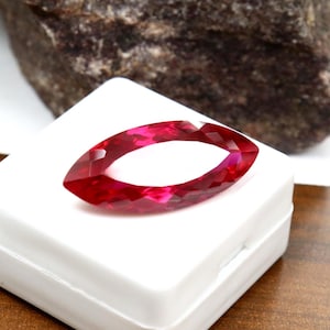 Könnte beinhalten: Ein Edelstein im Marquise-Schliff mit leuchtend roter, durchscheinender Farbe und facettierter Oberfläche. Der Stein liegt auf einer kleinen, quadratischen weißen Kunststoff-Präsentationsbox und zeigt seine längliche, spitz zulaufende Form.