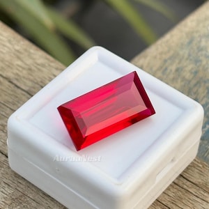 May include: A rectangular, faceted, deep red gemstone rests in a white plastic case. The gem is a vibrant, transparent red with sharp, clean lines. The case has the text "AuraaNest" printed on it.