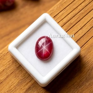 Pode incluir: Uma pedra preciosa de rubi estrela de cor vermelho profundo e formato oval, apresentando um padrão de estrela de seis pontas em sua superfície lisa e polida. A gema está colocada em uma caixa de exibição quadrada branca com o texto AuraaNest.