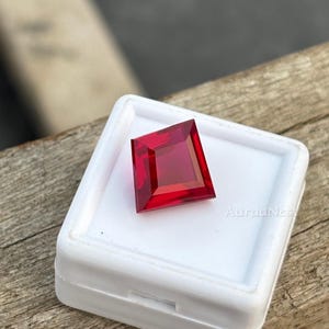 Puede incluir: Una gema roja intensa, tallada en forma cuadrada. La gema se presenta en una caja de plástico blanco, con el texto "AuraaNest" visible. La gema se exhibe sobre una superficie de madera.