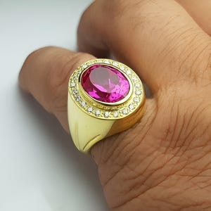 Op de afbeelding: Een gouden ring met een grote, ovale roze edelsteen. De edelsteen is omringd door kleinere, heldere stenen. De ring heeft een dikke, gepolijste gouden band, met een klassiek ontwerp.