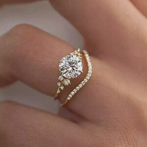 May include: A gold engagement ring set with a large, round, clear gemstone and a delicate band of smaller stones. The ring is set on a finger, showcasing its intricate design and sparkle. The band is a delicate gold color.