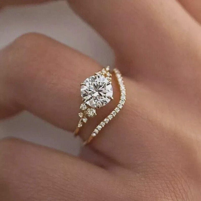 May include: A gold engagement ring set with a large, round, clear gemstone and a delicate band of smaller stones. The ring is set on a finger, showcasing its intricate design and sparkle. The band is a delicate gold color.