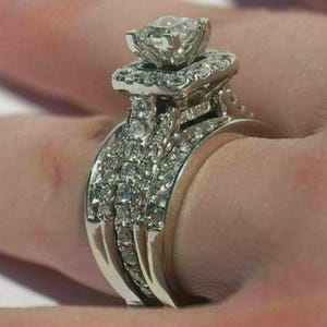 Pode incluir: Um anel de noivado de diamantes com uma pedra central de corte quadrado e um halo de diamantes menores. A banda também é incrustada com diamantes, criando um visual brilhante e luxuoso. O anel é feito de metal prateado.