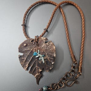 Puede incluir: Un collar artesanal con un colgante en forma de hoja de color bronce, adornado con piedras turquesas y verdes. Cuelga de un cordón de cuero trenzado marrón, con un cierre a juego. Una pieza de joyería única.
