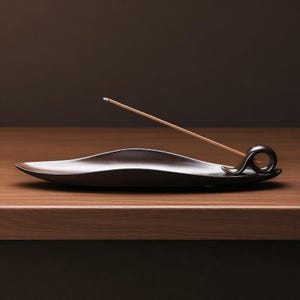 Könnte beinhalten: Ein dunkelbrauner, minimalistischer Räucherstäbchenhalter mit geschwungenem Design. Der Halter hat eine kleine Schlaufe an einem Ende, und ein schlanker Räucherstäbchen aus Holz ist darin platziert. Der Halter steht auf einer Holzoberfläche.