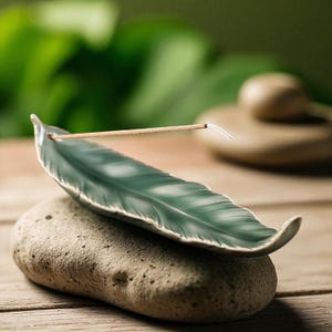 Könnte beinhalten: Ein teal-farbener Keramik-Räucherstäbchenhalter in Form einer Feder, der auf einem strukturierten grauen Stein liegt. Ein Räucherstäbchen steckt in der Halterung. Der Hintergrund ist mit grünem Laub verschwommen.