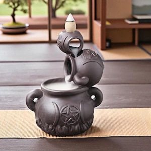 Puede incluir: Un quemador de incienso de cerámica marrón oscuro con un diseño en cascada. El quemador presenta una base de caldero, un recipiente intermedio y una pieza superior que sostiene un cono de incienso. El diseño incluye lunas crecientes y estrellas.