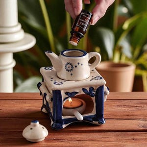 Peut inclure: Un diffuseur d'huiles essentielles en céramique blanc et bleu en forme de théière, avec un motif floral. Une bougie est allumée à l'intérieur. Une bouteille d'huile essentielle est versée. Posé sur une surface en bois.