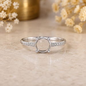 May include: A silver-coloured ring setting featuring a central four-prong mount designed to hold a round gemstone. The band is accented with a row of small, clear, round-cut stones embedded along each side of the shank, creating a polished finish.