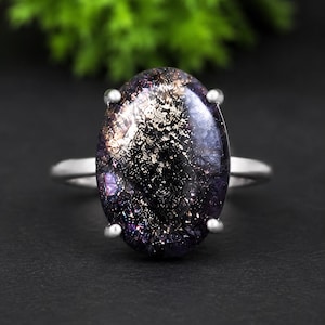 May include: A silver ring featuring a large, oval-shaped, dark purple gemstone with internal metallic, glitter-like inclusions. The stone is held in place by four silver prongs on a smooth, polished silver band, creating a reflective appearance.