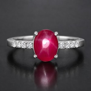 May include: A silver-toned ring featuring a central oval-shaped cabochon ruby with a visible star effect. The band is accented with three small, round, clear faceted stones on each side of the central gemstone, set in a polished metal frame.