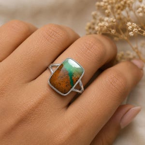 May include: A silver ring featuring a rectangular, polished gemstone with a distinct color split. The stone displays a textured, earthy brown section on the left and a smooth, vibrant mint green section on the right, set in a simple metal band.