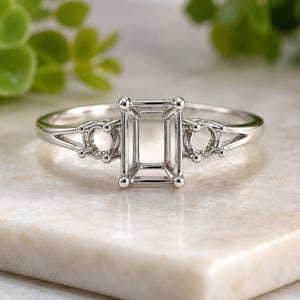 May include: A silver-toned metal ring setting featuring a central rectangular prong setting for an emerald-cut stone, flanked by two smaller circular prong settings on each side of the band, designed for a three-stone ring configuration.