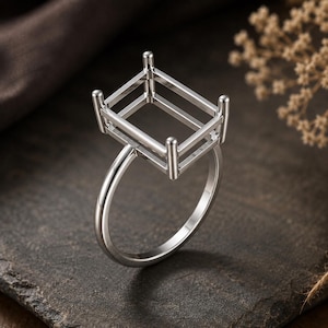 May include: A silver-toned metal ring setting featuring a rectangular basket head with four vertical prongs at the corners, designed to hold a gemstone. The band is smooth and circular, connecting to the base of the rectangular metal frame.