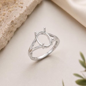 May include: A silver-toned metal ring setting featuring an oval-shaped prong mount designed to hold a gemstone. The band has a split-shank design that tapers toward the center, providing a polished, reflective surface and a structural frame for jewelry.