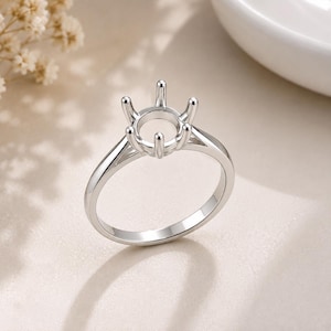 May include: A polished silver-colored metal ring setting featuring a six-prong head designed to hold a single gemstone. The band is smooth and tapers slightly toward the top, connecting to a circular gallery that supports the prong structure.