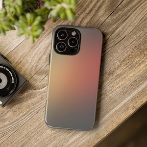 May include: A smartphone case with a gradient design, shifting from yellow to pink and then to grey. The case has a glossy finish and a camera cutout with multiple lenses. The phone case is on a wooden surface.