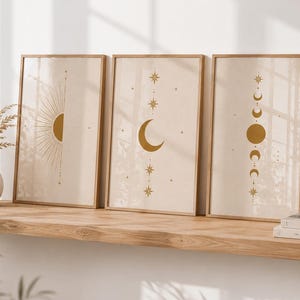 May include: Three rectangular framed wall art prints featuring gold celestial motifs on a light beige background. The left print shows a sunburst, the centre shows a crescent moon, and the right shows a vertical sequence of moon phases.