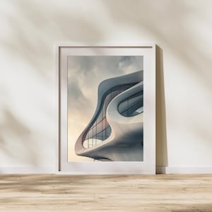 May include: Framed architectural print featuring a modern building with a curved, white facade and large glass windows. The building's design is organic and flowing, set against a cloudy sky. The print is in a white frame, displayed on a wooden floor.