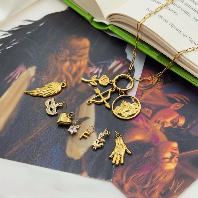 May include: A collection of gold-colored charms and a necklace. Charms include a wing, a number eight, a heart, a flower, the letter "F", a horseshoe, a hand with an eye, a rose, and an arrow and bow. The necklace has a mountain pendant.