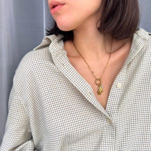 May include: A gold-colored necklace with a charm shaped like a hand holding a small, clear stone. The necklace is worn over a plaid button-down shirt in shades of white, beige, and navy blue.