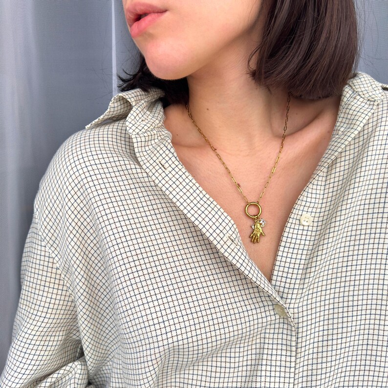 May include: A gold-colored necklace with a charm shaped like a hand holding a small, clear stone. The necklace is worn over a plaid button-down shirt in shades of white, beige, and navy blue.