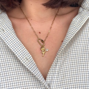 May include: Gold-toned necklace featuring a rose pendant and a small heart-shaped charm. The necklace has a delicate chain and a carabiner-style clasp. The rose pendant is detailed with intricate petals and leaves.