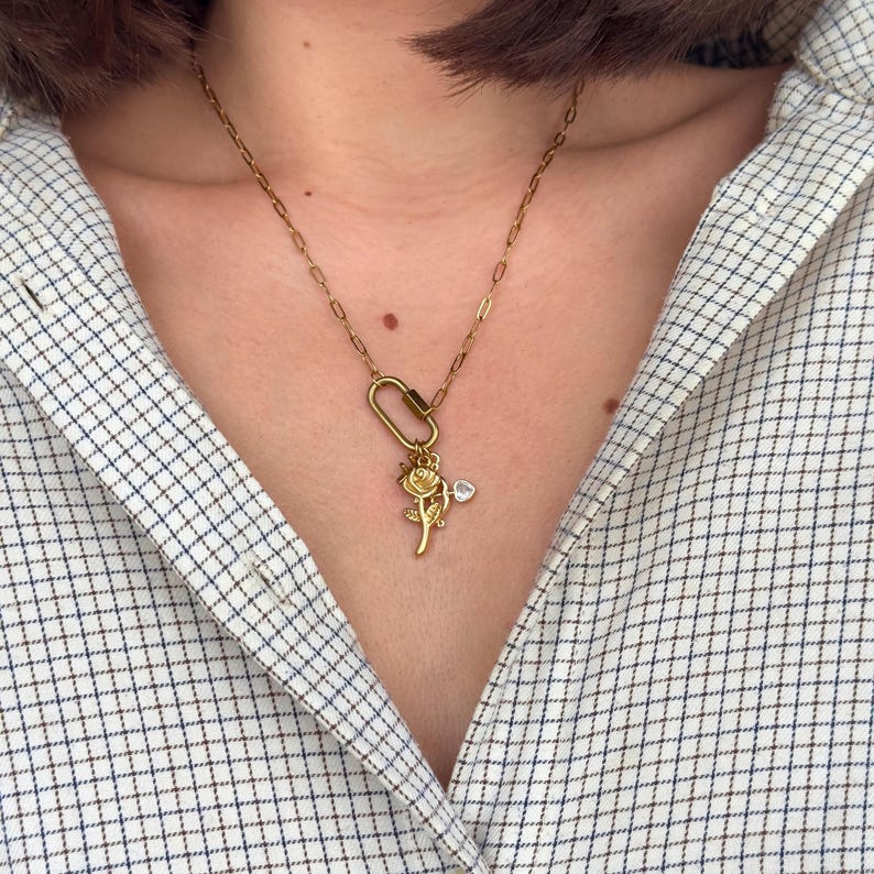 May include: Gold-toned necklace featuring a rose pendant and a small heart-shaped charm. The necklace has a delicate chain and a carabiner-style clasp. The rose pendant is detailed with intricate petals and leaves.