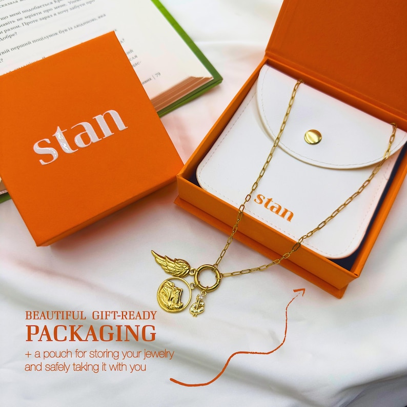 May include: An orange gift box and a white pouch with a gold necklace. The necklace has a gold chain and three charms: a wing, a coin, and a small anchor. The box and pouch have the word "stan" printed on them.