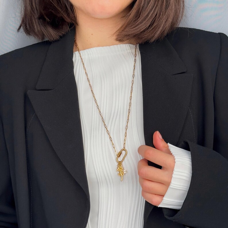 May include: A gold-colored necklace with a rose pendant and a clasp. The necklace is worn over a white ribbed top and a black blazer. The rose pendant has a small crystal accent.