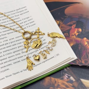May include: Gold-toned necklace with various charms, including a heart, a hand with an eye, a rose, a bow and arrow, and a wing. The charms are arranged on a delicate chain, creating a layered effect. The necklace is displayed on a book.