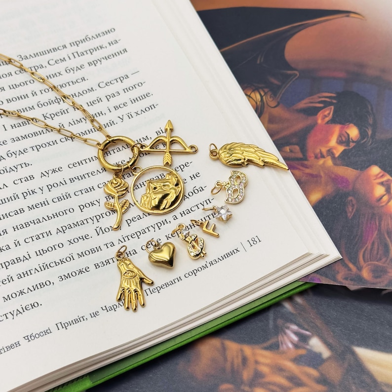 May include: Gold-toned necklace with various charms, including a heart, a hand with an eye, a rose, a bow and arrow, and a wing. The charms are arranged on a delicate chain, creating a layered effect. The necklace is displayed on a book.