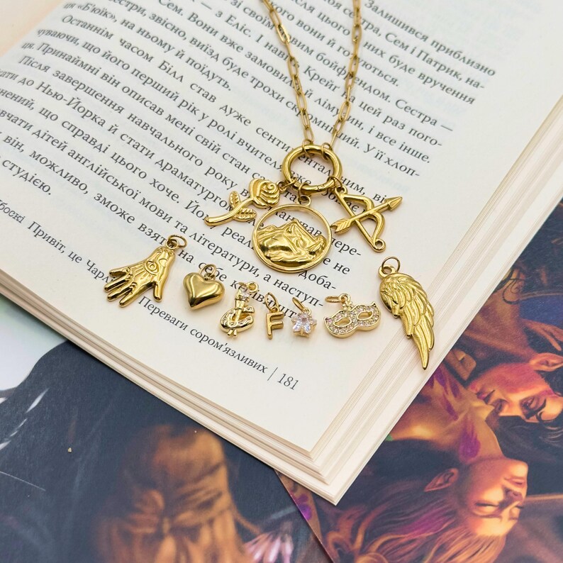 May include: A gold-colored necklace with multiple charms, including a rose, mountain scene, bow and arrow, hand, heart, crown, letter F, flower, mask, and wing. The necklace is displayed on an open book.