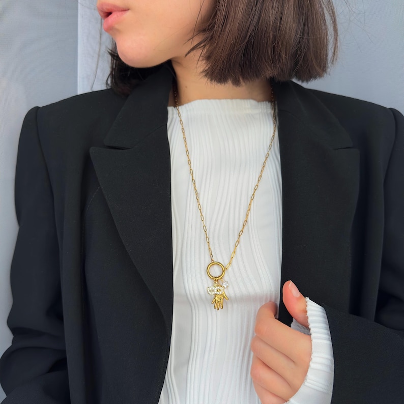 May include: A gold-colored necklace with a pendant featuring a hand and a ring. The necklace is worn over a white ribbed shirt and a black blazer. The pendant has small, clear embellishments.