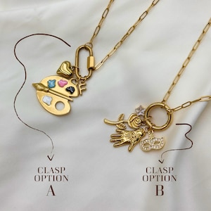May include: Two gold-colored necklaces with different charm options. One features a painter's palette with colorful paint, a heart, and the letter "F". The other has a rose, hand, mask, and a star-shaped charm. The text reads "Clasp Option A" and "Clasp Option B".