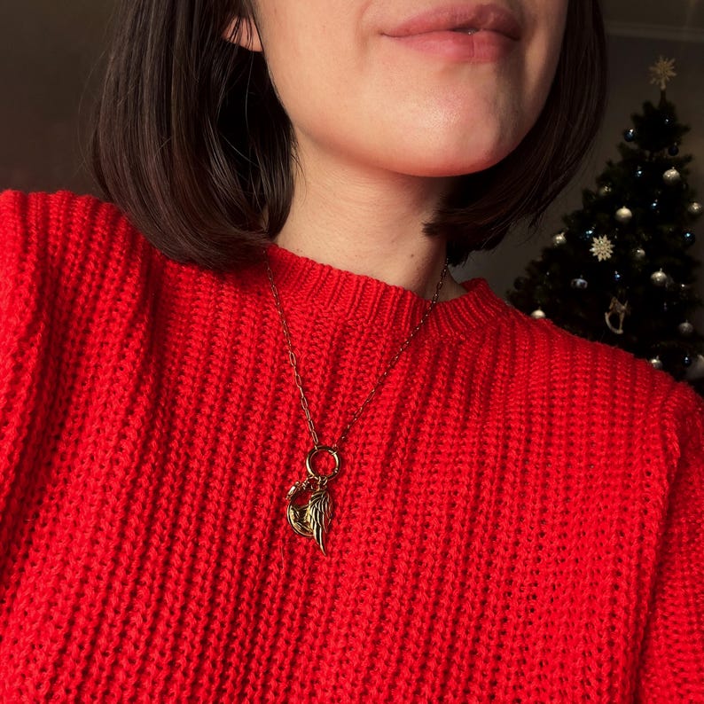 May include: A gold-colored necklace with a pendant featuring a heart and a wing design. The necklace is worn over a bright red sweater. A Christmas tree is visible in the background.