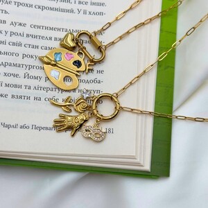 May include: Two gold-colored necklaces with unique charms. One features a palette with colorful paint, a heart, and a key. The other has a hand, a rose, a mask, and a small charm with the number 181. The necklaces are displayed on an open book.